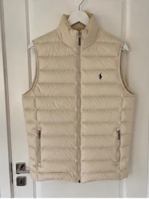 Polo Ralph Lauren Performance väst – Beige/Sand – Nyskick - Polo Ralph Lauren Performance-väst i beige/sand. Väldigt stilren och lätt att matcha, med klassisk Polo Pony-logga på bröstet, quiltad design, ståkrage och dragkedjefickor.  Västen är i nyskick och endast använd ett fåtal gånger. Inga hål, fläckar eller andra skador. Fyllningen är fortfarande väldigt fin och västen är väl omhändertagen.  Jag köpte den själv på Farfetch för några månader sedan. Originaletiketten med Polo Ralph Lauren. Pris kan naturligtvis diskuteras 