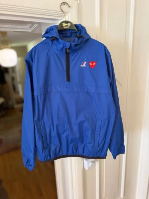 Blå vindjacka från K-Way x Comme des Garçons - Snygg blå vindjacka från K-Way i samarbete med Comme des Garçons Play. Jackan har huva med dragsko, halv dragkedja framtill och elastiska muddar. På bröstet finns K-Way-logga och det ikoniska röda hjärtat med ögon. Perfekt för blåsiga dagar. Köpt på NK i GBG! 