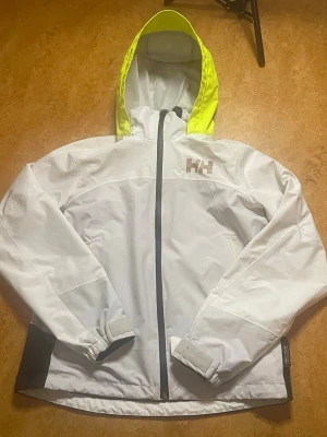 Vit regnjacka från Helly Hansen - Snygg vit regnjacka från Helly Hansen med svart dragkedja och detaljer i neongult på huvan. Jackan har justerbara muddar, två sidofickor och HH-logga på bröstet. Perfekt för regniga dagar och outdoor-äventyr.