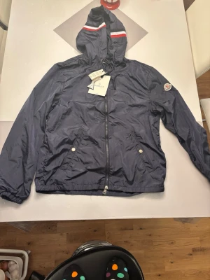 Marinblå vindjacka från Moncler - Snygg marinblå vindjacka från Moncler med huva och vita samt röda detaljer på huvan. Jackan har dragkedja framtill, två fickor med tryckknappar och Moncler-logga på ärmen. Perfekt för blåsiga dagar och enkel att matcha till olika outfits.