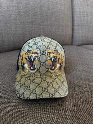 Gucci keps  - Snygg keps från Gucci med klassiskt GG-mönster i beige och svart. Framtill finns två stora, färgstarka tigrar. Baksidan är i svart mesh för extra ventilation. Perfekt accessoar för dig som vill sticka ut med en exklusiv look.