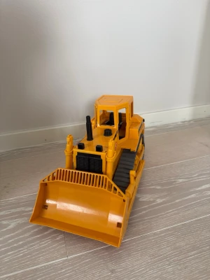 Gul leksaksbandtraktor - Säljer en gul leksaksbandtraktor i plast med detaljer som hytt, larvband och stor skopa framtill. Perfekt för dig som gillar byggfordon och vill ha något kul att leka med inomhus eller utomhus.