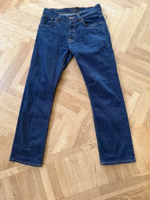 Nudie Jeans Average Joe Dry Denim - Snygga mörkblå jeans med en klassisk fem ficksmodell. Jeansen har kontrastsömmar och är tillverkade i ett robust denimtyg. Nudie Jeans Average Joe Dry denim i Storleken 32/34, med måtten 40 i midja och 101 på längden! Stor potential för fina fades och slitningar i framtiden!