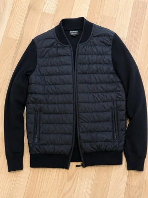 Svart cardigan från barbour - Snygg svart bomberjacka från Barbour med quiltad front och släta ärmar. Jackan har ribbade muddar, dragkedja framtill och två fickor med dragkedja. Klassisk collegekrage och stilren design som passar till det mesta.