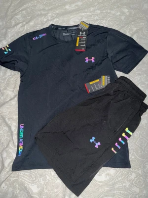 Svart träningsset från Under Armour - Säljer ett svart träningsset från Under Armour med t-shirt och shorts. Setet har färgglada loggor och detaljer i regnbågsfärger. Materialet är lätt och andas, perfekt för träning eller gym. T-shirten har kort ärm och rund hals, shortsen har resår i midjan.