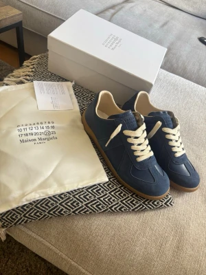 Maison Margiela blå sneakers - Snygga blå sneakers från Maison Margiela med beige detaljer och vita skosnören. Ovandelen är i mocka och läder, och sulan är i gummi med klassiskt mönster. Perfekta för dig som gillar stilrena och trendiga skor.