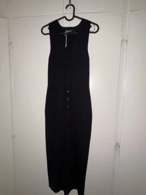 Svart långklänning från Gina Tricot - Snygg svart långklänning från Gina Tricot med v-ringning och knappar längs hela framsidan. Klänningen är ärmlös och har en smickrande, rak passform. Perfekt att styla med sneakers eller boots för en trendig look.