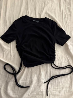 Svart croppad topp med snörning - Snygg svart croppad topp med korta ärmar och trendig snörning i sidan som ger en cool rynkad effekt. Tillverkad i ett mjukt, ribbat material som sitter bekvämt. Något nopprig men väldigt enkelt att få bort!! 
