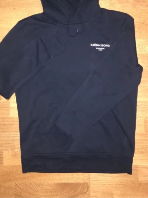 Svart hoodie från Björn Borg - Svart hoodie från Björn Borg med vit logga på bröstet. Klassisk modell med huva och känguruficka framtill. Mjuk bomullskänsla och ribbade muddar vid ärmslut och nederkant. Perfekt för en avslappnad och stilren look.
