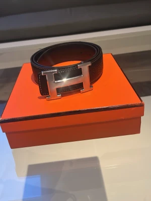 Svart bälte med silverfärgat H-spänne - Snyggt svart bälte med ett stort, blankt H-format spänne i silverfärg. Bältet har en stilren och klassisk design som passar till många olika outfits. Tillverkat i skinn och levereras i en orange ask. ( får med box )