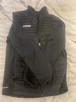 Svart vindjacka från CCM - Snygg svart vindjacka från CCM med quiltade partier på bröst och sidor. Jackan har hel dragkedja framtill, hög krage och diskret logga på bröstet. Materialet är lätt och vindtätt, perfekt för blåsiga dagar. Sportig och stilren design.