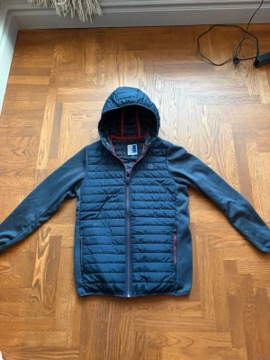 Mörkblå hybrid jacka från Jack & Jones  - Snygg mörkblå vindjacka med quiltad front och huva. Jackan har dragkedja framtill, två fickor med röda detaljer och mjuka ärmar i sweatshirtmaterial. Perfekt för blåsiga dagar och enkel att matcha till olika outfits. Storlek 164. Aldrig använd
