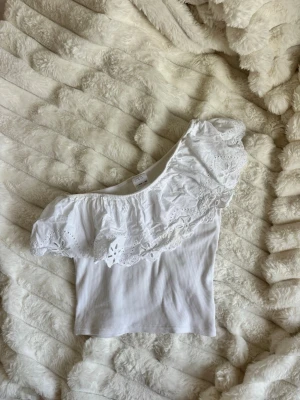Vit oneshoulder topp - Säljer en vit oneshoulder topp med bred spetsvolang och ribbad struktur. Toppen är ärmlös på ena sidan och har en romantisk känsla med fina broderade detaljer i volangen. Perfekt för dig som vill sticka ut lite extra.