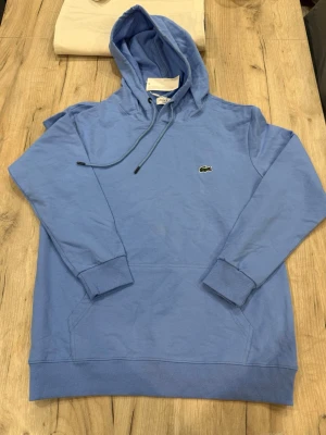 Blå hoodie från Lacoste - Snygg ljusblå hoodie från Lacoste med klassisk krokodillogo på bröstet. Regular fit med huva och dragsnören, samt ribbade muddar vid ärmar och nederkant. Perfekt för en chill och stilren look. Tillverkad i mjukt bomullsmaterial.