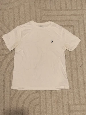 Vit t-shirt från Polo Ralph Lauren - Klassisk vit t-shirt från Polo Ralph Lauren med den ikoniska broderade loggan på bröstet. T-shirten har rund hals och korta ärmar, tillverkad i mjuk bomull för en skön känsla. Perfekt basplagg till garderoben. Stl 150!