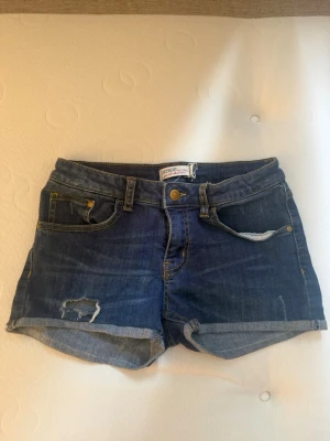 Blå denimshorts från Detroit - Snygga blå denimshorts från Detroit med upprullade benslut och slitna detaljer framtill. Klassisk femficksmodell med bälteshällor och knappstängning. Perfekta för varma dagar och enkel att matcha med allt.