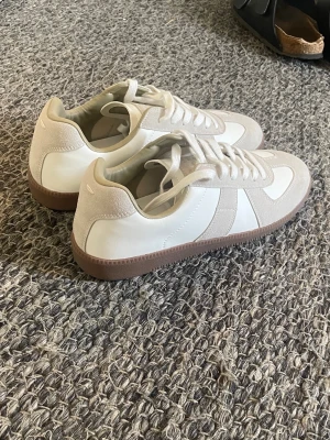 Maison Margiela gata  - Säljer ett par stilrena vita sneakers med beige mockadetaljer och bruna gummisulor. Skorna har klassisk låg profil och snörning. Perfekta för dig som gillar minimalistisk och clean stil. Materialet är en mix av läder och mocka.
