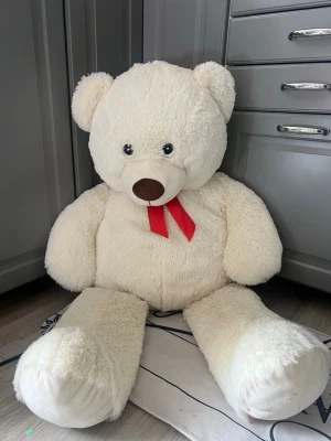 Stor vit nallebjörn med röd rosett - Jättestor mjuk nallebjörn i vit plysch med en söt röd rosett runt halsen. Perfekt som mysig inredningsdetalj eller present. Nallen har stora svarta ögon och en brun nos. Sitter fint på golvet och är supergosig. 