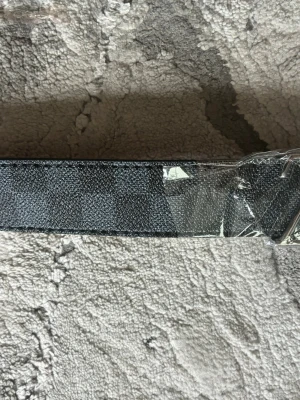 Svart och grått rutigt bälte - Snyggt bälte i svart och grått med rutigt mönster och metallspänne. Bältet har flera hål för justering och en stilren design som passar till många olika outfits. 116cm