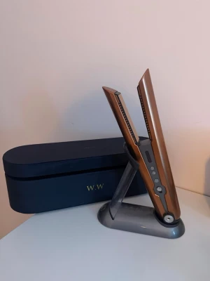 Dyson kopparfärgad plattång - Snygg och modern plattång från Dyson i kopparfärg med svarta och silverdetaljer. Kommer med ett matchande svart fodral och laddningsställ. Perfekt för att styla håret med sladdlös frihet och avancerad teknik. Helt oanvänd 
