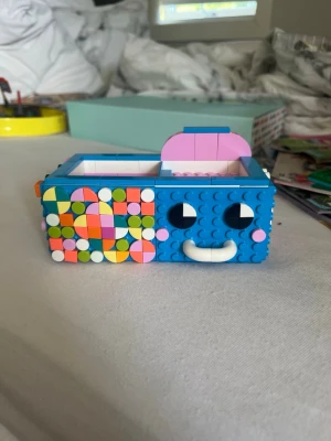 Färgglad LEGO-förvaringslåda - Säljer en unik förvaringslåda byggd av LEGO med blå front, glada ansiktsdetaljer och färgglada mönster på sidorna. Lådan har två fack och en utdragbar låda, perfekt för småprylar eller smycken. Materialet är plast och designen är lekfull och kreativ. De kommer isär och men massor av färgglada plupparna så man kan designa hur man vill