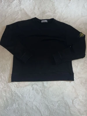 Svart sweatshirt från Stone Island - Klassisk svart sweatshirt från Stone Island med rund halsringning och ikonisk logopatch på vänster ärm. Tröjan har ribbade muddar vid ärmslut och nederkant. Perfekt för en stilren och avslappnad look.