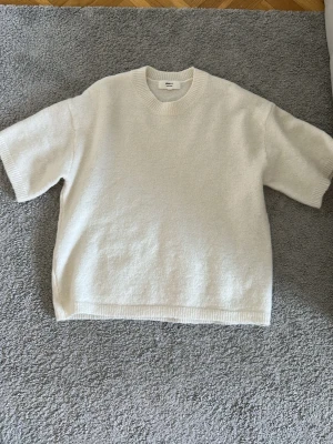 Vit stickad t-shirt med kort ärm - Säljer en stilren vit stickad t-shirt med korta ärmar och rund hals. Tröjan har en avslappnad passform och ribbade kanter vid hals, ärmslut och nederkant. Perfekt för lager på lager och enkel att matcha med olika outfits.