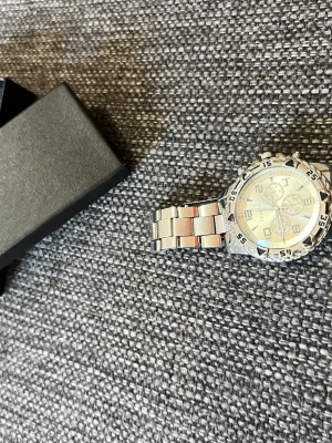 Silverfärgad kronografklocka i metall - Stilren silverfärgad kronografklocka med länkarmband i metall. Klockan har en rund urtavla med tydliga siffror och markeringar, samt flera små urtavlor för extra funktioner. Perfekt accessoar för dig som gillar en sportig men ändå klassisk look.