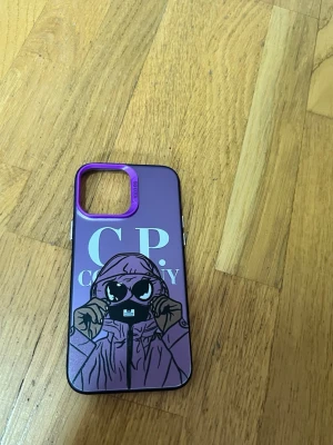 Lila mobilskal med CP Company-motiv - Snyggt mobilskal i lila med motiv av en figur i hoodie och stora glasögon samt texten 'C.P. Company'. Skalet har en markerad lila ram runt kameran och är gjort i hårdplast. Perfekt för dig som vill ha ett unikt och trendigt skydd till mobilen.