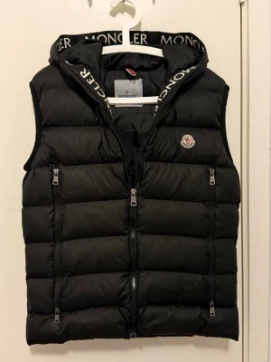 Svart Moncler   - Snygg svart dunväst med avtagbar huva och dragkedja framtill. Västen har två sidofickor med dragkedja och en bröstficka med Moncler-logga. Tjockt vadderad för extra värme och sportig look. Perfekt för lager-på-lager-stil.