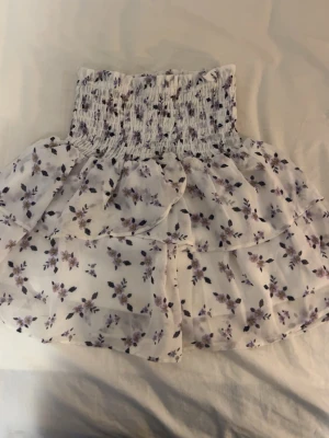Vit blommig kortkjol med volang - Supersöt vit kortkjol med lila och svarta blommor. Kjolen har smock i midjan och dubbla volanger som ger en luftig och lekfull känsla. Perfekt för varma dagar och enkel att matcha med en enkel topp.