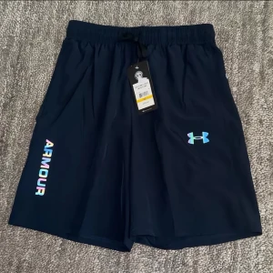 Mörkblå shorts från Under Armour- M junior. S Herr - Snygga mörkblå shorts från Under Armour med logga och texten 'ARMOUR' i regnbågsfärger på ena benet. Shortsen har elastisk midja med snörning och är perfekta för träning eller chill. Lätta och bekväma med sportig känsla.                              Passform: M men kan passa herr som bär lite mindre strl alltså S för herr