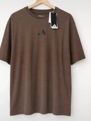 Ny brun Adidas t-shirt med logga - Säljer en brun t-shirt från Adidas med liten logga i leopardmönster på bröstet och tryck på ryggen. T-shirten har rund hals, korta ärmar och en relaxed passform. Perfekt för dig som gillar streetwear och vill ha något unikt i garderoben.