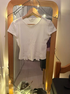  T-shirt - Säljer en vit t-shirt med korta ärmar och rund hals. T-shirten har två dekorativa knyt framtill vid nederkanten som ger en unik touch. Tillverkad i mjukt bomullsmaterial, perfekt för en avslappnad och stilren look.