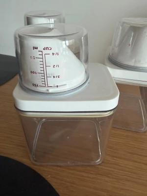 Set med förvaringsburkar och mått - Säljer ett set med tre genomskinliga förvaringsburkar i plast med vita lock. På varje lock sitter ett mått i plast, perfekt för att mäta ingredienser eller tvättmedel. Burkarna är fyrkantiga och stapelbara, smidiga för köket.