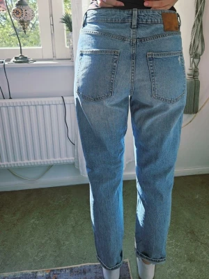 157 blå jeans st M - Säljer ett par ljusblå mom jeans med hög midja och raka ben. Jeansen har slitningar vid knäna och klassiska fem fickor. Materialet är denim i bomull och passformen är avslappnad och trendig.