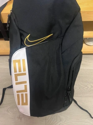 Nike Elite svart ryggsäck - Snygg svart Nike Elite ryggsäck med gulddetaljer och vit panel. Ryggsäcken har flera fack, vadderade axelband med genomskinliga detaljer och tydliga Nike-loggor. Perfekt för träning eller skola, rymmer mycket och har sportig design.