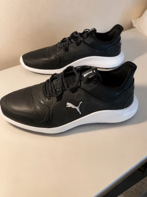 Svarta Puma sneakers med vit sula - Snygga svarta sneakers från Puma med vit sula och svartvita skosnören. Skorna har en stilren design med Pumas logga på sidan och ovandel i syntetmaterial. Perfekta för dig som gillar sportig och clean stil.