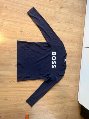 Mörkblå långärmad t-shirt med BOSS-tryck - Mörkblå långärmad topp med stort vitt BOSS-tryck på bröstet. Klassisk rak passform och rund halsringning. Tillverkad i mjuk bomull för en bekväm känsla.