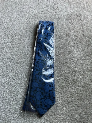 Blå slips med paisleymönster - Snygg blå slips med paisleymönster i olika nyanser av blått. Slipsen har en klassisk form och är tillverkad i ett glansigt tyg som ger en elegant känsla. Perfekt för att lyfta din outfit med lite extra stil.