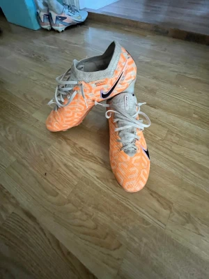 Nike fotbollsskor air zoom - Säljer ett par grymma fotbollsskor från Nike i orange och beige med vita snören och svart swoosh på sidan. Skorna har en stickad ovandel för skön passform och är perfekta för dig som vill ha kontroll på planen. Snygg design som sticker ut på fotbollsplanen!