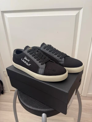 Svarta Saint Laurent sneakers - Snygga svarta sneakers från Saint Laurent med vit sula och svart textilovandel. Klassisk låg modell med svarta skosnören och Saint Laurent-logga broderad i vitt på sidan. Perfekta för en stilren och trendig look.