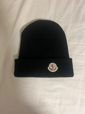 Svart ribbstickad mössa Moncler - Snygg svart ribbstickad mössa från Moncler med uppvikt kant och klassisk Moncler-logga framtill. Perfekt för dig som vill ha en stilren och varm accessoar till vintern. Materialet är mjukt och stretchigt för skön passform.