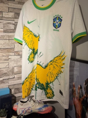 Vit Brasilien fotbollströja Nike - Snygg vit fotbollströja från Nike med Brasilien-emblem och gröna samt gula detaljer. Framsidan har ett stort grafiskt tryck av en gul fågel och gröna accenter på krage och ärmslut. Perfekt för dig som älskar fotboll och vill sticka ut på planen.