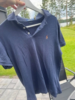 Marinblå piké från Polo Ralph Lauren - Klassisk marinblå pikétröja från Polo Ralph Lauren i modellen Custom Slim Fit. Tröjan har korta ärmar, krage och knappar framtill samt den ikoniska broderade loggan på bröstet. Tillverkad i mjuk bomull för en bekväm känsla.