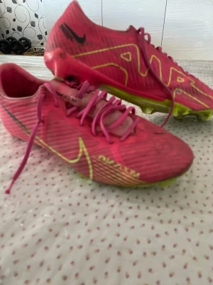 Nike Air Zoom rosa fotbollsskor - Säljer ett par Nike Air Zoom fotbollsskor i en riktigt snygg rosa färg med gula och svarta detaljer. Skorna har snörning, låg profil och dobbar för bra grepp på planen. Tillverkade i syntetmaterial för lätt känsla och bra bollkontroll.