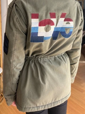 Olivgrön parkajacka med broderi - Snygg olivgrön parkajacka med broderad text 'Love' i rött, vitt och blått på ryggen från märket Zadig Voltaire. Jackan har fyra stora fickor framtill, dragsko i midjan och patch på ärmen. Perfekt för dig som vill ha en unik och trendig jacka. 💕