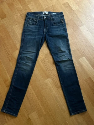 Mörkblå slim fit jeans - Säljer ett par mörkblå Replay Anbass slim fit jeans med snygga slitningar och klassiska fem fickor. Jeansen har kontrastsömmar och en läderpatch bak i midjan. 