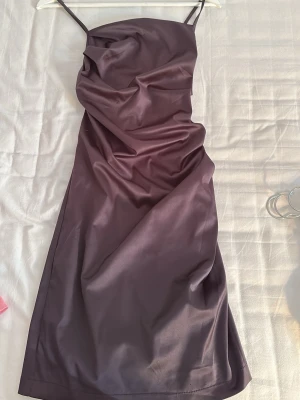 Mörklila satin klänning med smala band - Säljer en elegant mörklila klänning i satin med smala axelband och draperad framsida. Klänningen har en lång siluett och justerbara band i ryggen för perfekt passform. Perfekt för dig som vill ha en stilren och trendig look.
