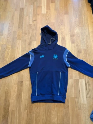 Blå hoodie från Puma Marseille - Snygg marinblå hoodie från Puma med Olympique de Marseille-logga på bröstet. Hoodien har ljusblå detaljer på axlarna, vit piping och en stor ficka framtill. Perfekt för dig som gillar sportig stil och vill representera ditt favoritlag.